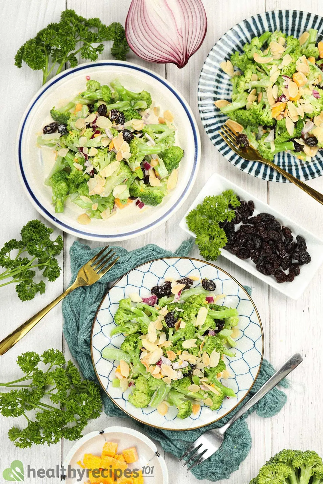 A high-angle shot of three broccoli salad plates, decorated by a blue mesh cloth, a fork, a plate of cheddar cubes, raisins, fresh parsley, and broccoli heads