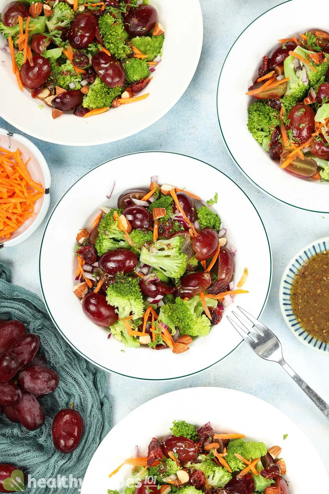  A close-up photo of plates of colorful salad with roasted broccoli, cherry tomatoes, red onions, and almonds, drizzled with a zesty dressing. The salad is placed on a wooden table with a fork on the side.