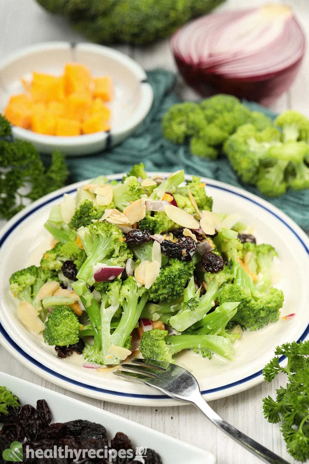 A white plate with blue rim with broccoli florets, raisins, sliced almonds, and a fork laid on it