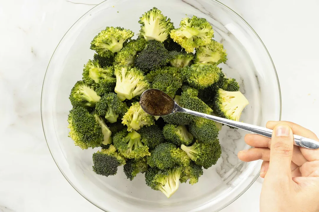 A hand distributing a brown dressing into a bowl of broccoli florets.