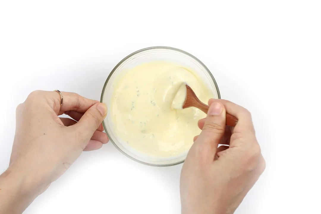 A hand using a wooden spoon to mix a thick, pale yellow sauce.