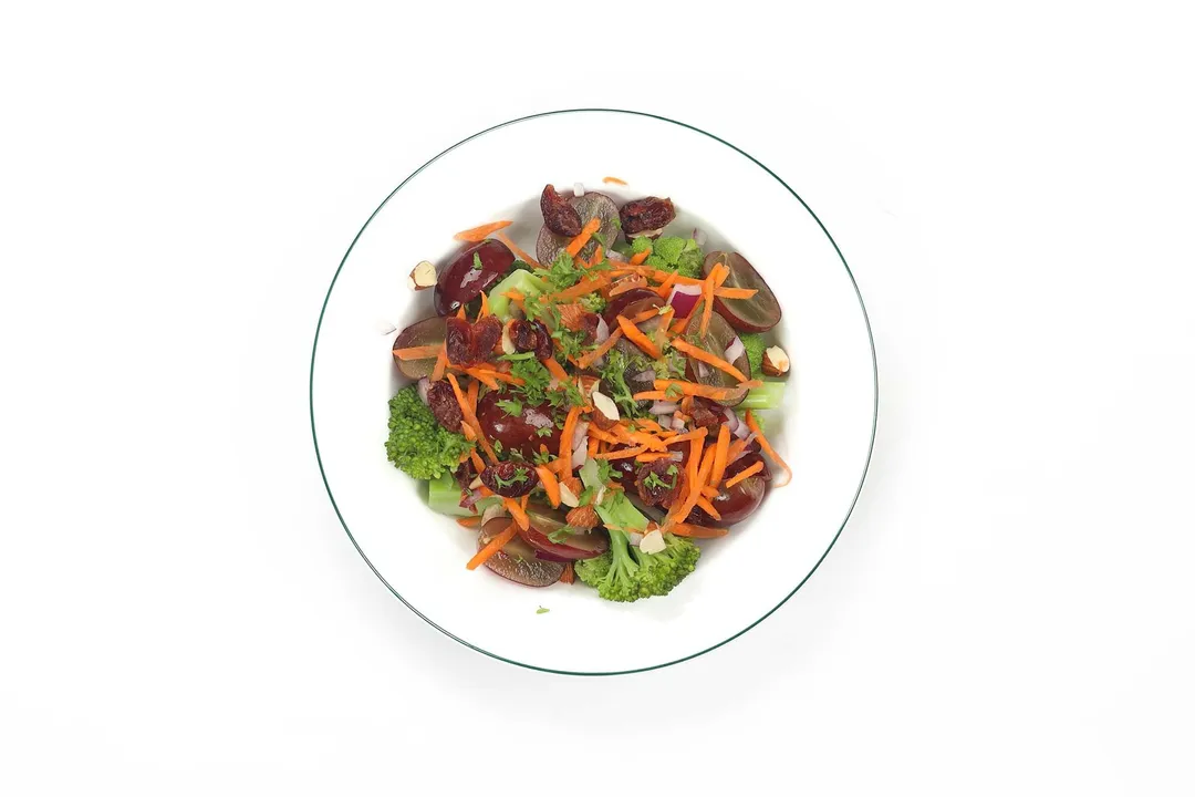 A white plate filled with broccoli florets, red grapes, and julienned carrrot strips.