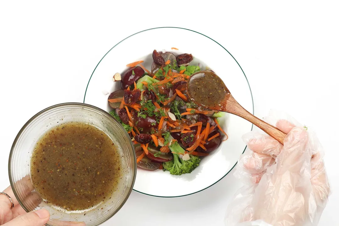 A hand using a wooden spoon to distribute a dark brown dressing onto a plate of vegan broccoli salad.