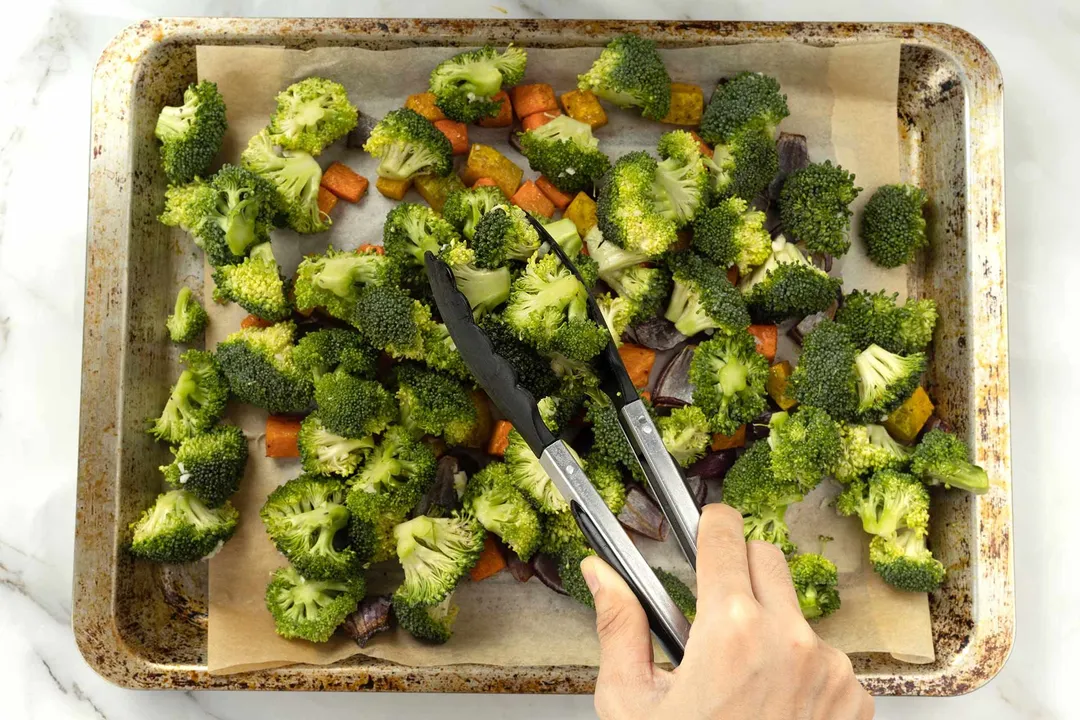 A hand using a tong to stir broccoli florets and squash cubes spread out on a baking sheet lined with parchment paper.