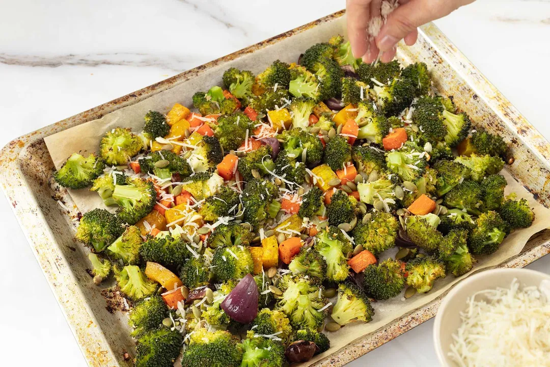 A hand sprinkling shredded cheese onto a sheetpan filled with broccoli florets and squash cubes.