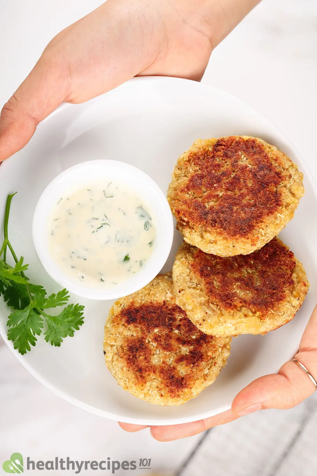 two hand holds a white plate with three cauliflower patties and a small bowl of sauce