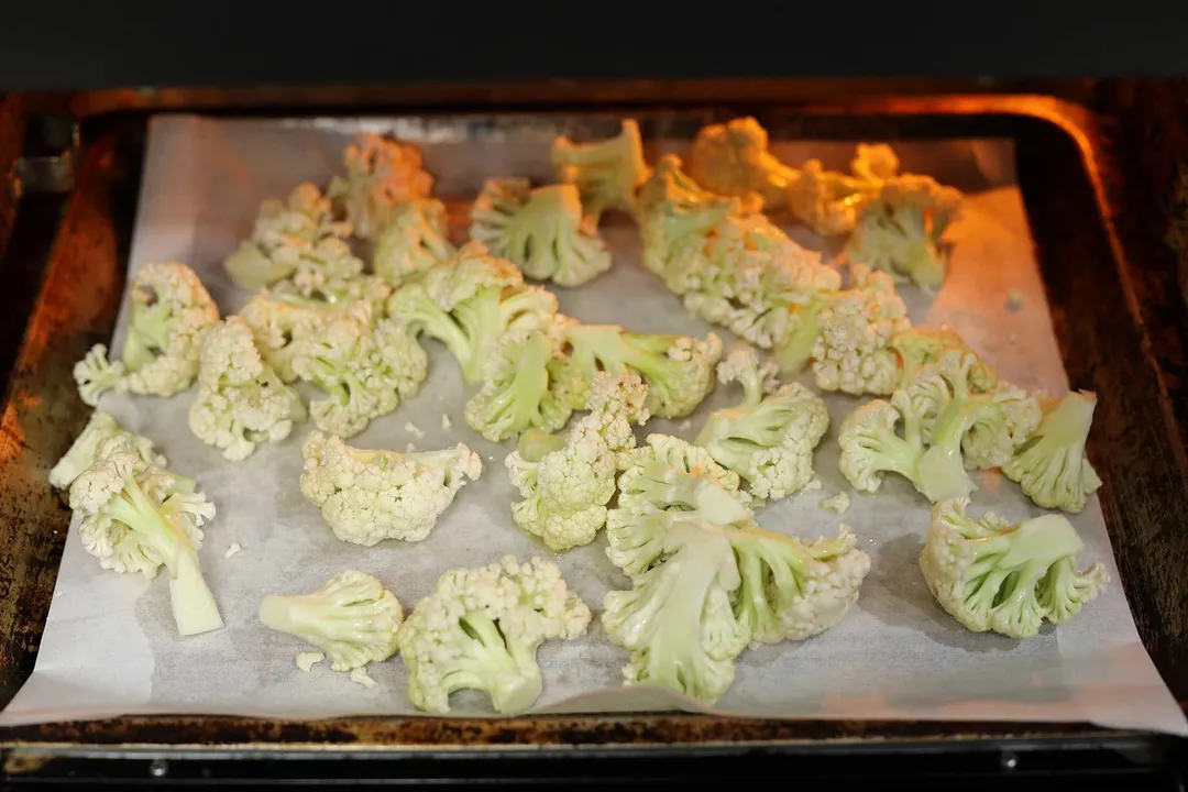 A baking sheet lined with parchment paper and filled with cauliflower florets cooking inside an oven