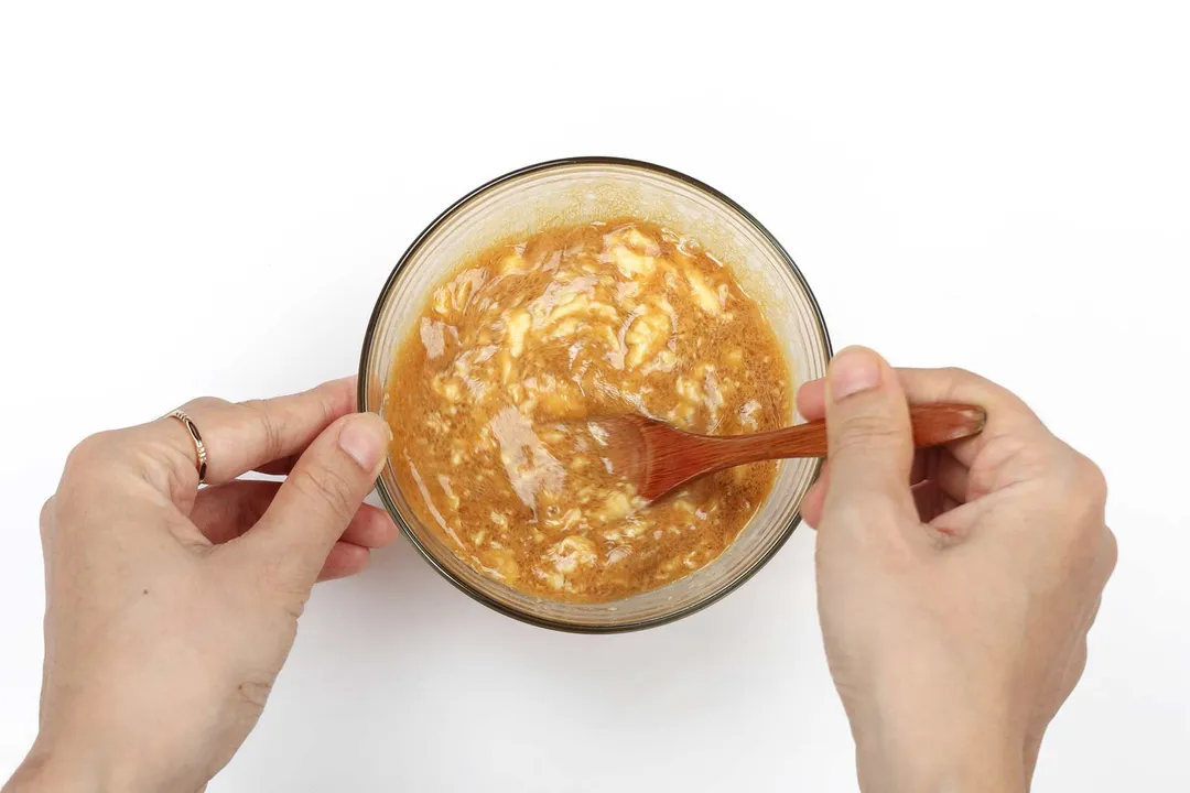 Two hands stirring caramel brown sauce with white stripes in a small glass bowl with a wooden spoon