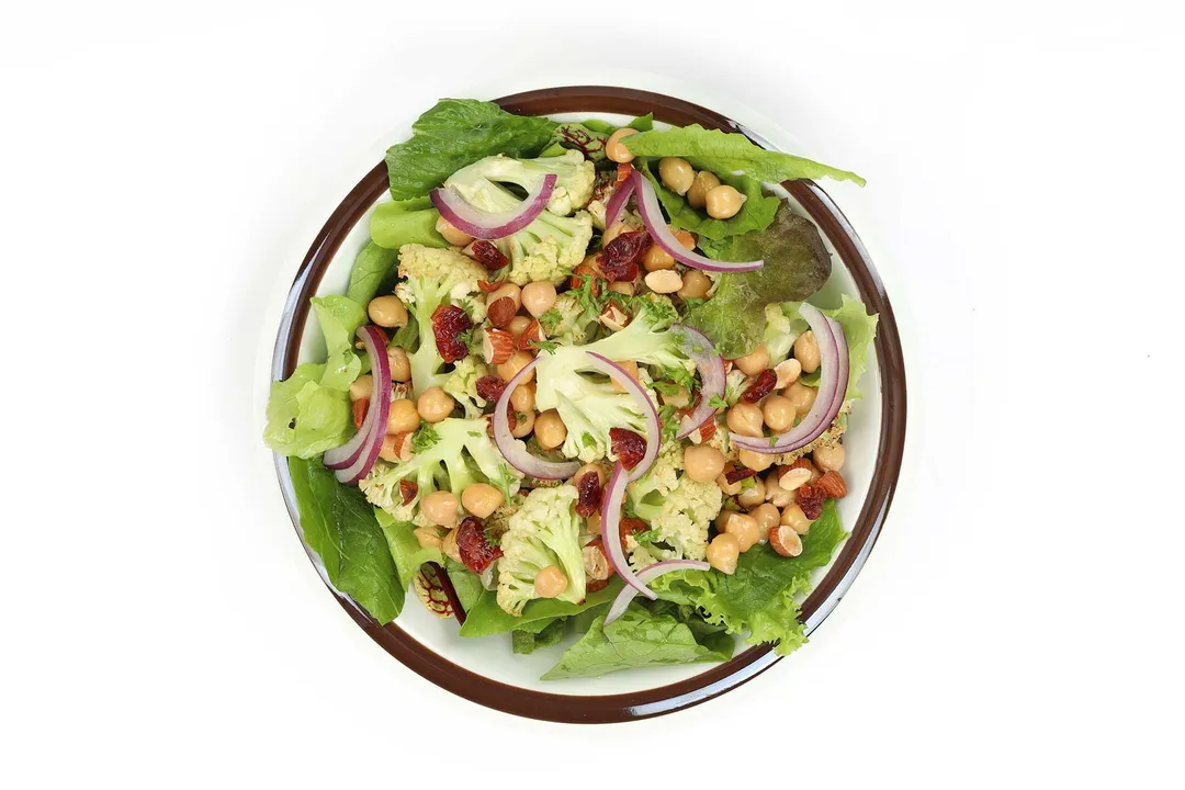 A white round plate with brown rim containing a bed of lettuce leaves filled with chickpeas, sliced red onion, dried cranberries, and cauliflower florets