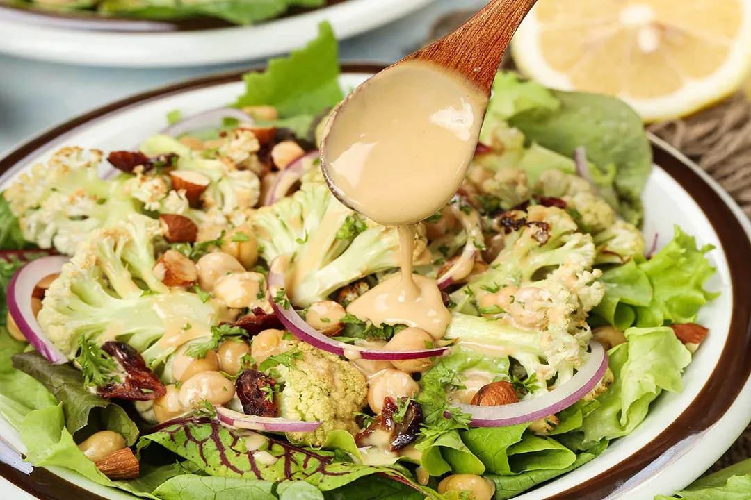 A wooden spoon drizzling a pale orange and thick sauce onto a plate of roasted cauliflower salad