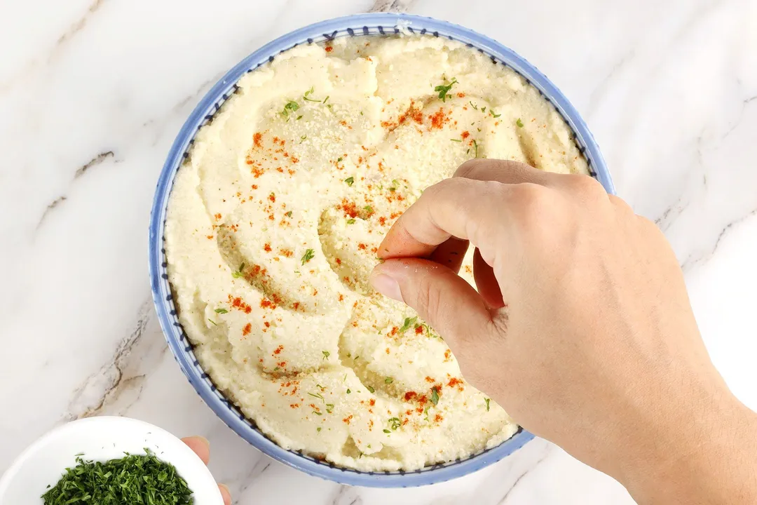 hand sprinkle parsley on top of a bowl of mashed cauliflower