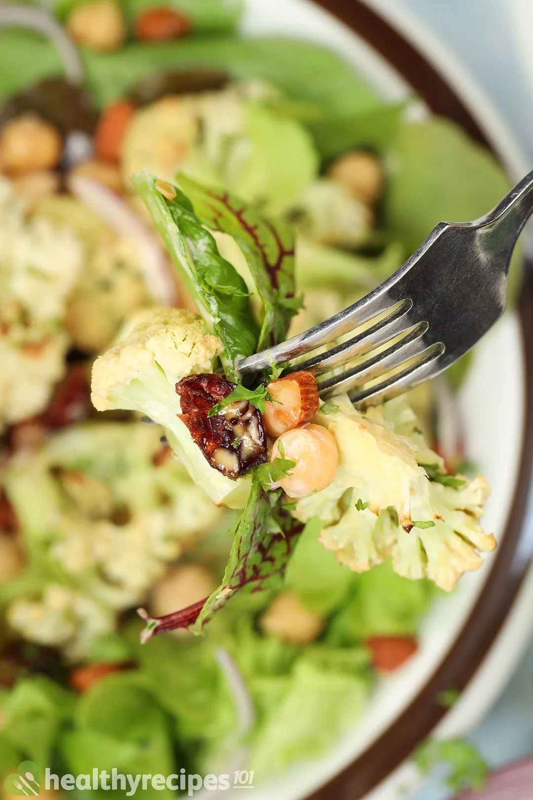 A fork piercing into a cauliflower floret, a lettuce leave, a chickpea, half a walnut, and a dried cranberry