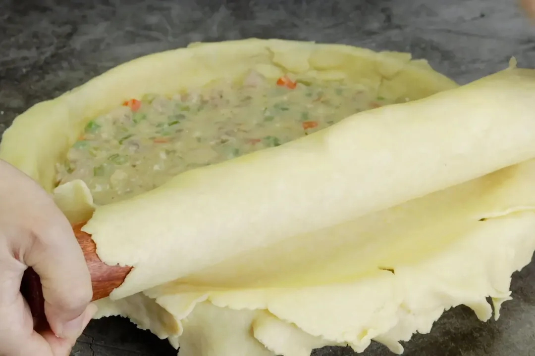 A hand rolling the dough over the surface of a food bowl while making chicken pot pie