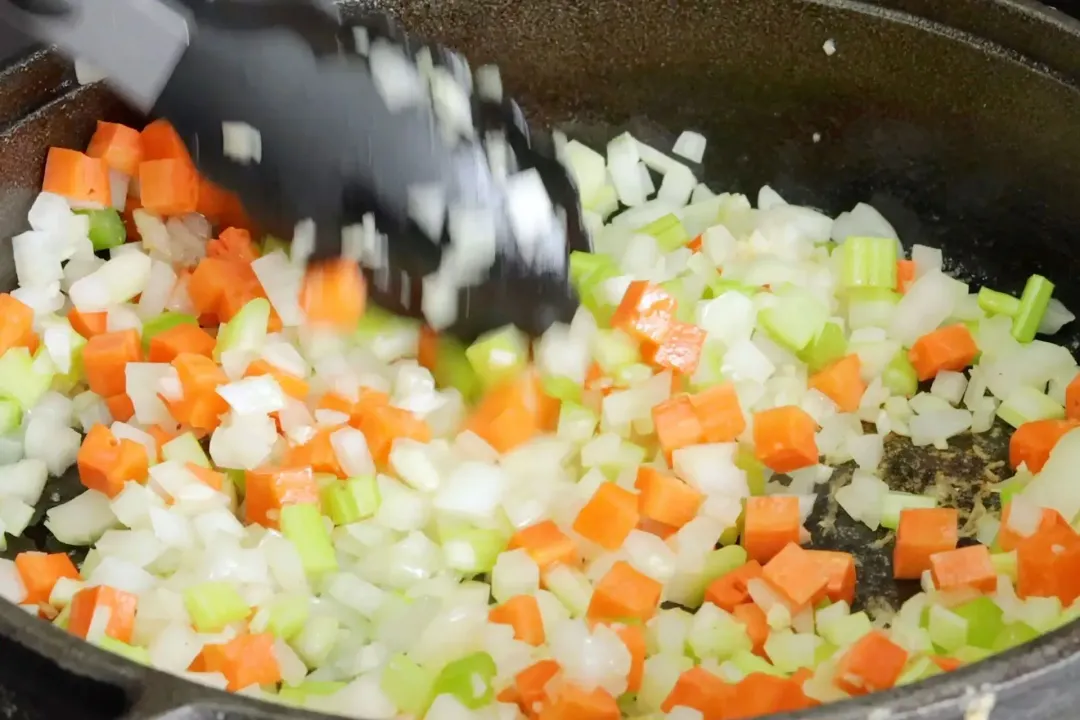 A pair of tongs used to saute diced carrots, diced onion, and diced celery in a pot.