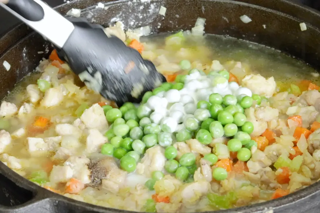 A pair of tongs mixing green peas with diced vegetables in a broth soup pot