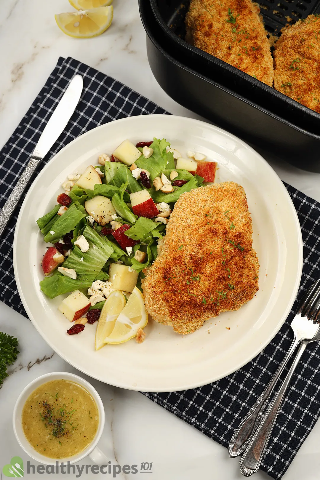 a plate of cooked chicken breast and salad next to an air fryer basket and a small bowl of sauce