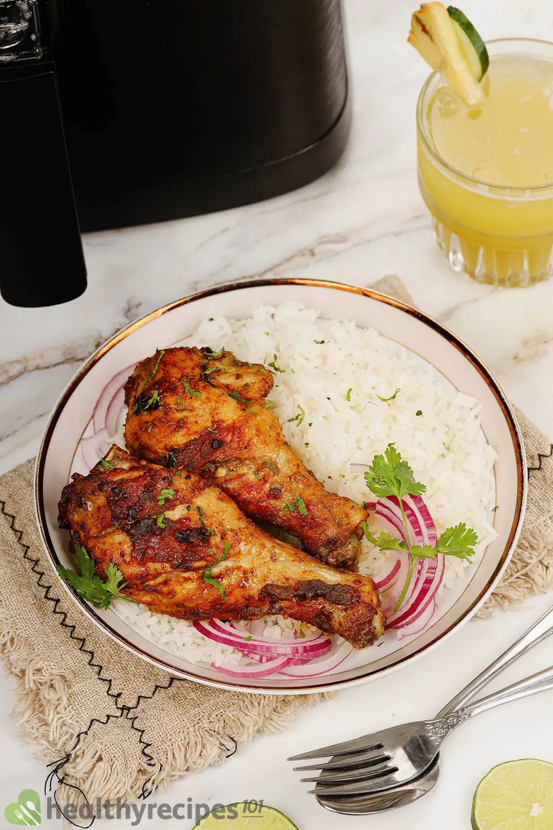 two chicken drumsticks bed on rice and slices onion on a plate decorated with a glass of juice and a spoon, a fork next to an air fryer