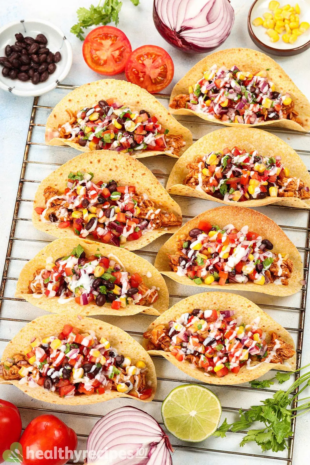 8 BBQ chicken tacos laid on a baking rack and surrounded by fresh coriander, half a lime, red onion halves, tomato halves, and a small dish of red beans