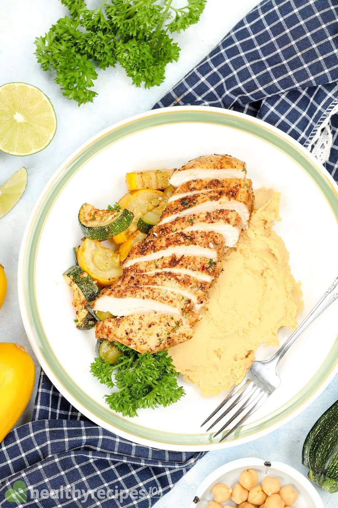 A spiced and cut-up chicken breast served with potato mash, grilled zucchini rounds, parsley on a white plate with fork, on a checkered blue cloth