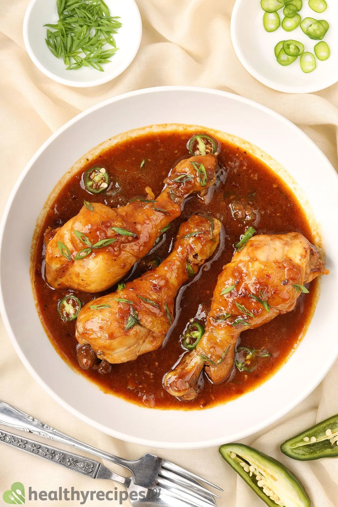 a white plate of three cooked chicken legs with sauce, garnished with chopped scallion, sliced pepper