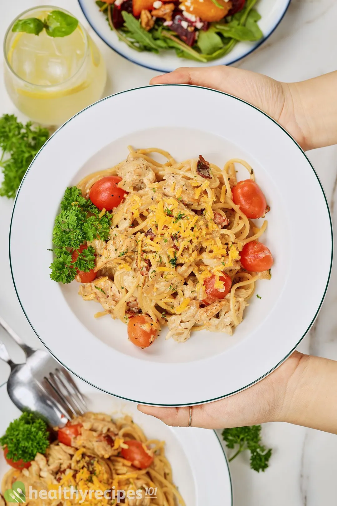 two hand holding a plate of cooked chicken with pasta and half cherry tomatoes, garnished with parsley