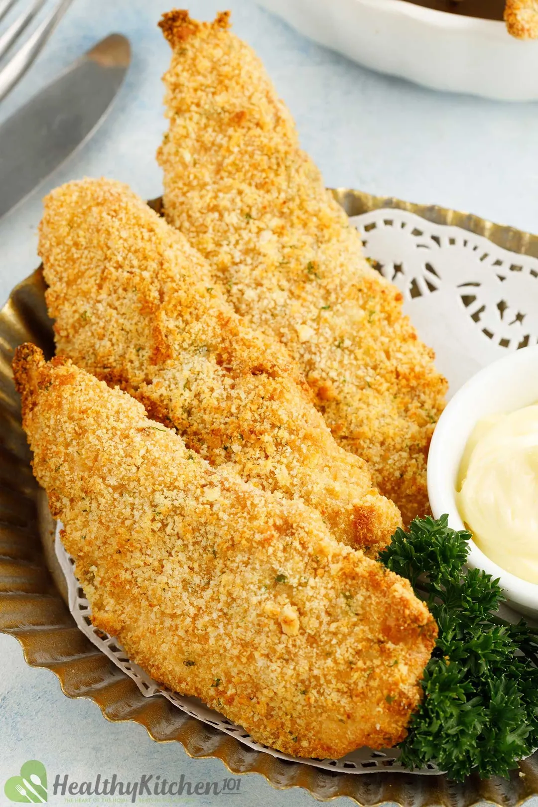 three cooked chicken tenderloin on a plate with a small bowl of white sauce and parsley for garnish