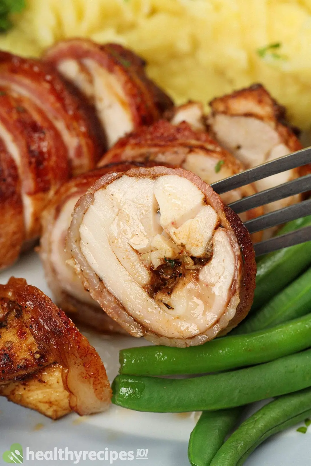 close-up shot of sliced chicken wrapped by bacon and holding by a fork, decorated with green beans, mashed potatoes