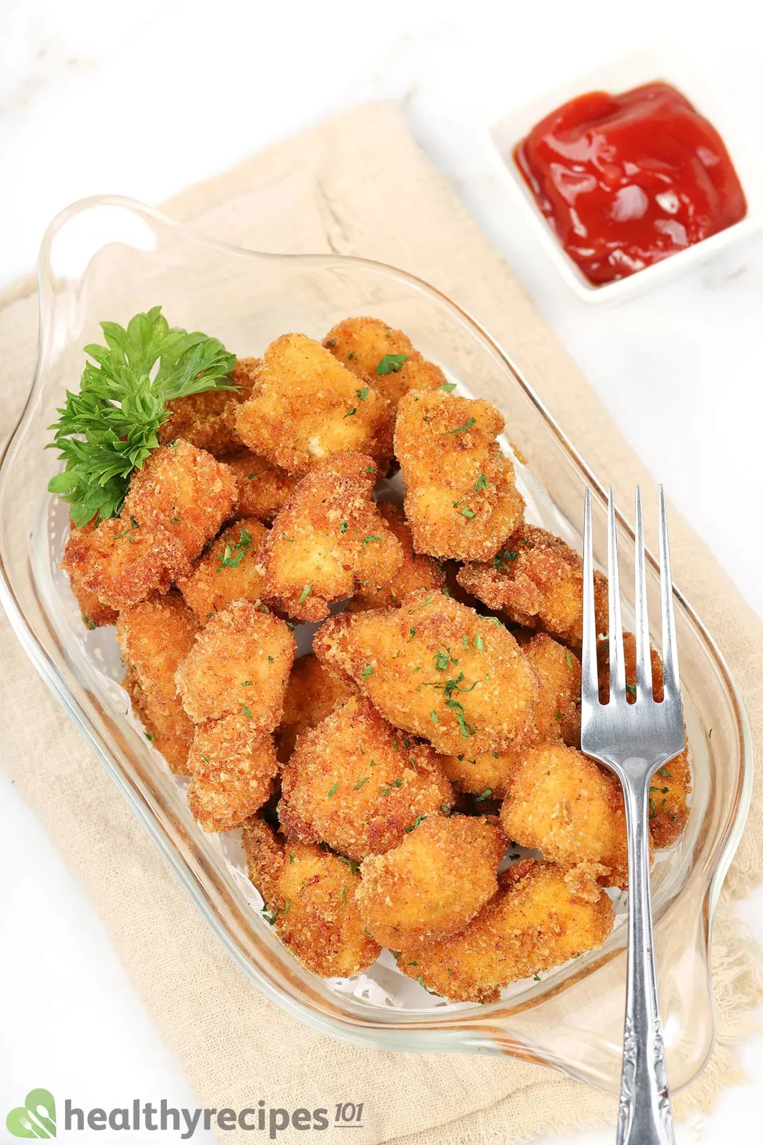 cooked chicken cubed on a glass tray decorated with a fork, a small bowl of ketchup and parsley