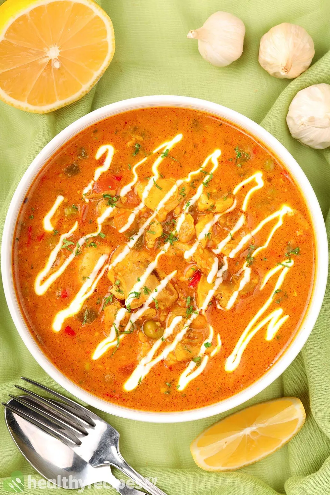 top view of a bowl of cooked chicken with orange sauce and top with yogurt, surrounded by spoon and fork, half lemon, garlic