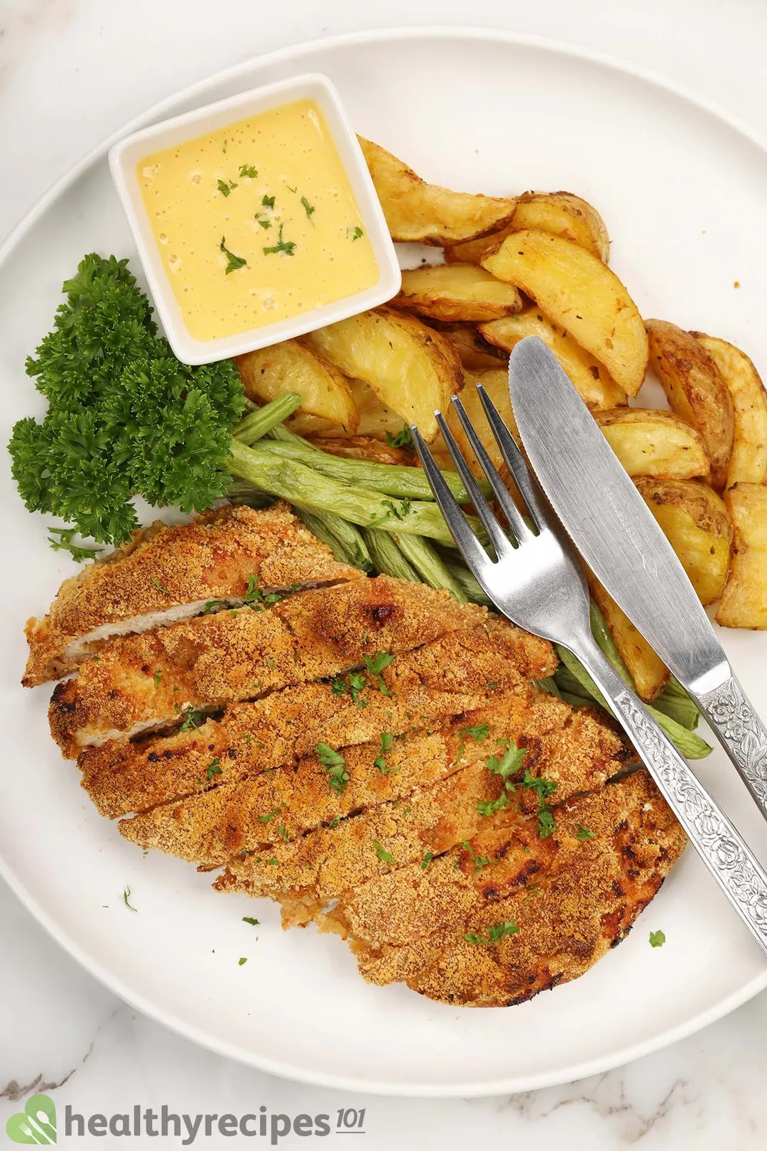 a plate of cooked boneless chicken breast with potato wedge and green bean next to a small bowl of yellow sauce decorated with parsley and a fork, a spoon