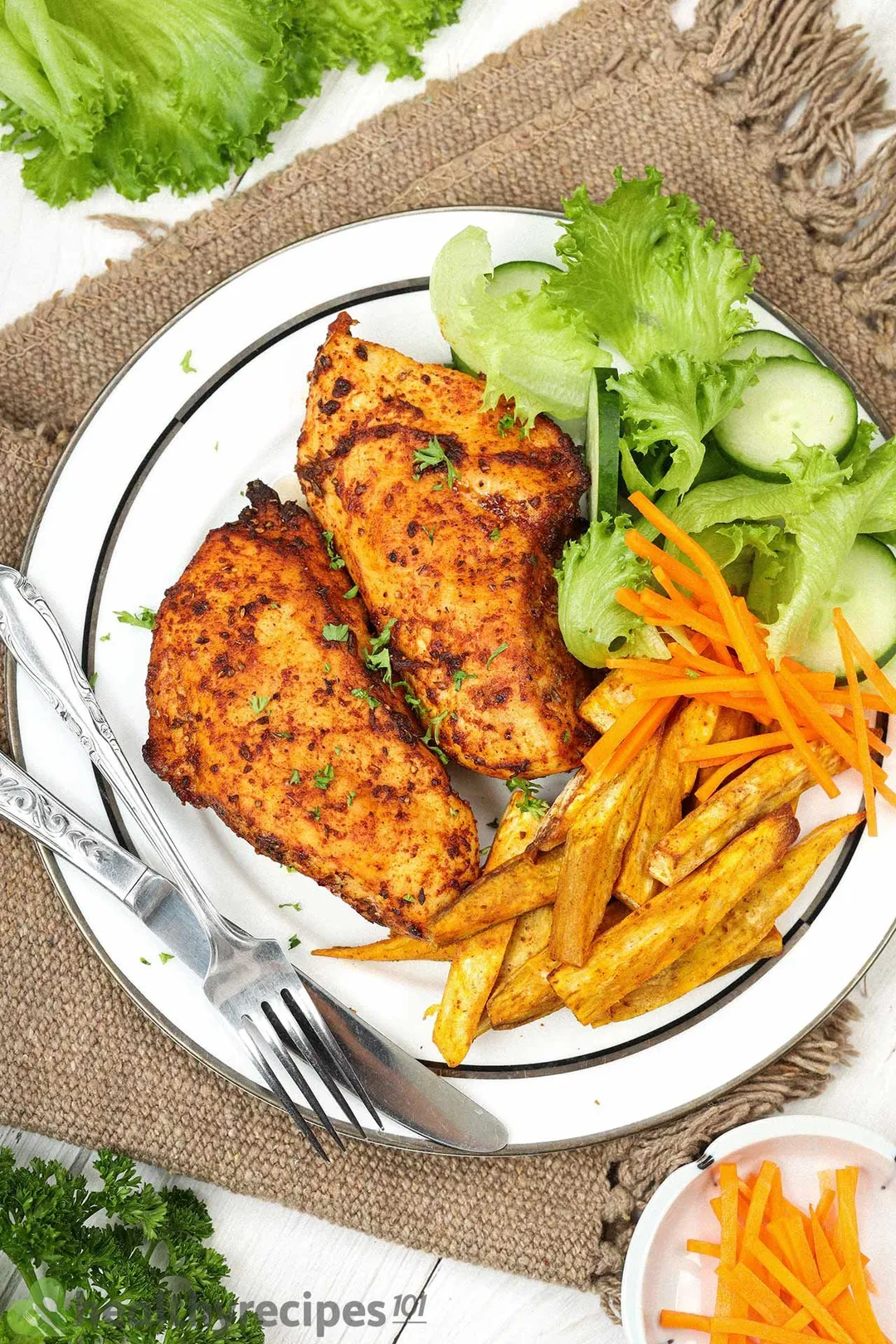 A plate of cooked chicken breasts covered in spices, fried sweet potatoes, lettuce, and cucumber slices