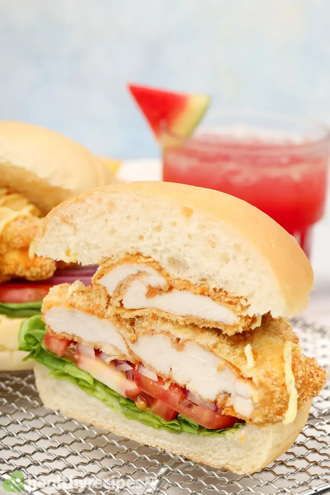 Half a chicken burger with lettuce, tomatoes, and deep-fried chicken breasts in between buns on a wire rack next to a glass of watermelon juice