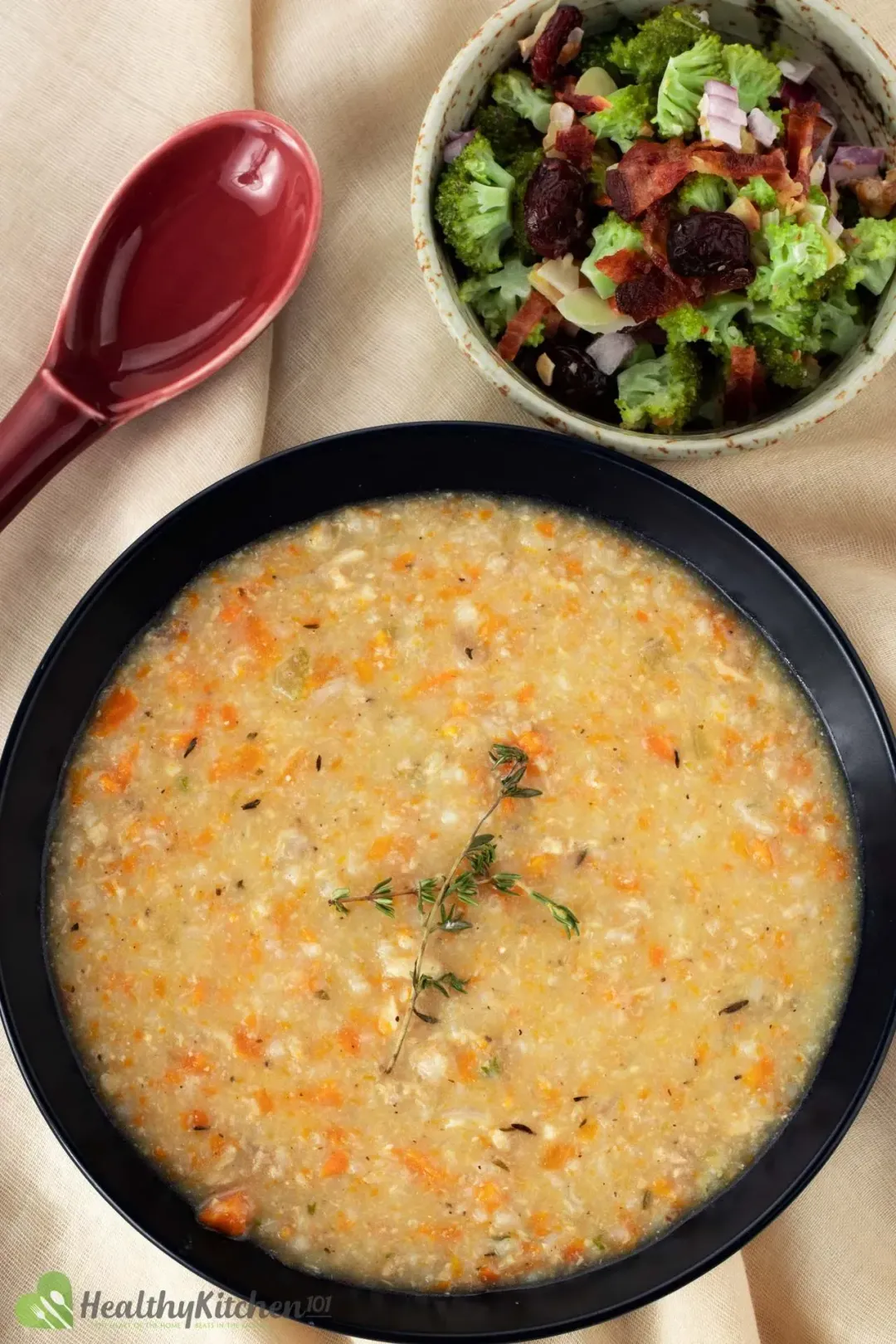 A bowl of chicken and rice soup topped with two sprigs of rosemary next to a salad and a red spoon