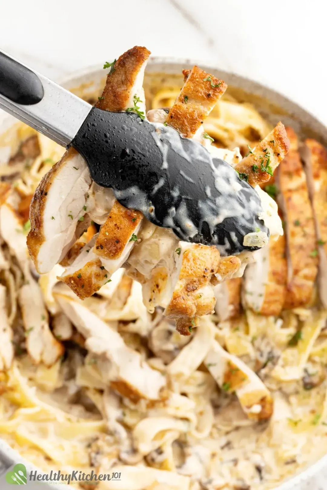 Tongs picking up slices of cooked chicken from a pan of Chicken Alfredo