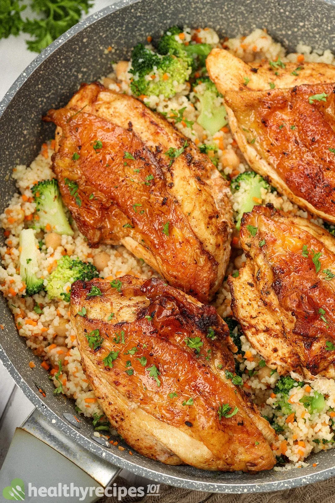 A close-up shot of a large and deep pan filled with cooked couscous, broccoli florets, chickpeas, and browned chicken thighs