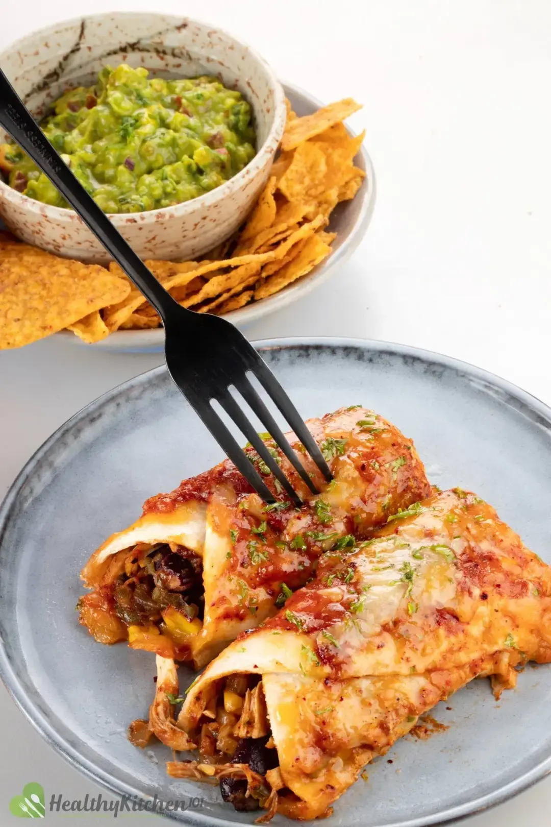 Two enchilada rolls on a blue plate with a black fork dunked in, with some tortillas and guacamole in the back