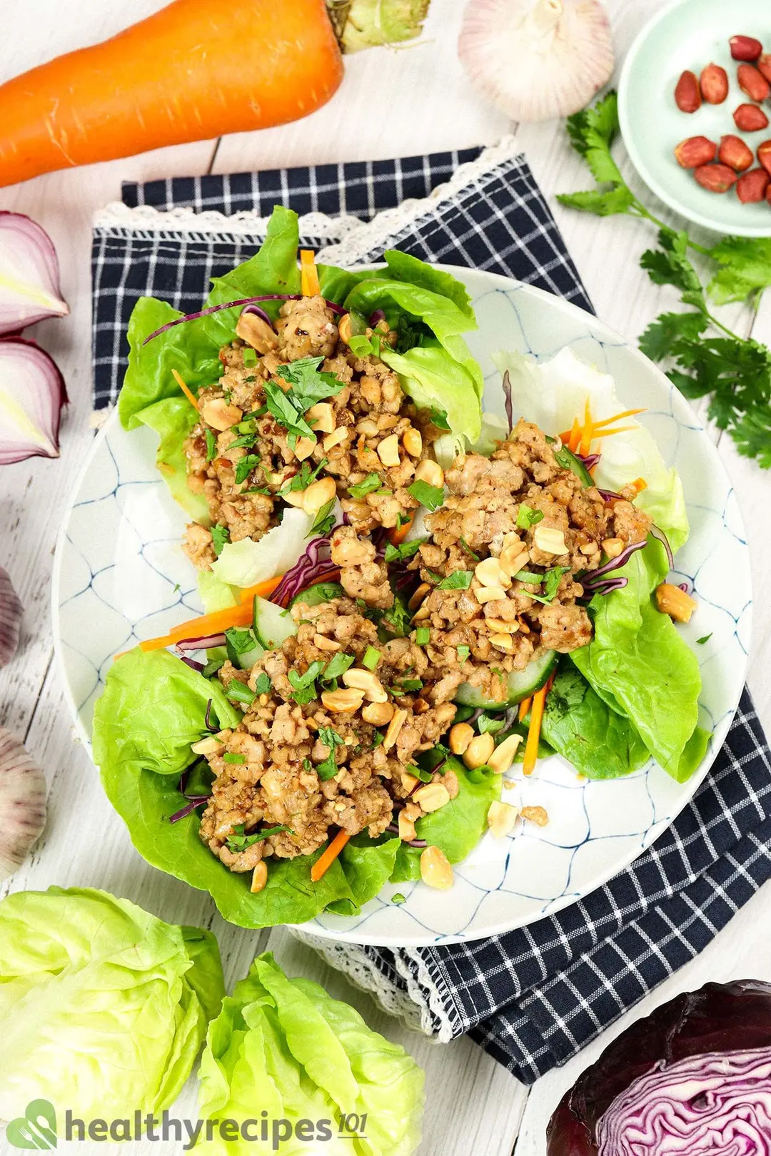 A dish of lettuce wraps placed on a folded tablecloth and surrounded by a carrot, lettuce heads, half a red cabbage, red onion halves, and coriander