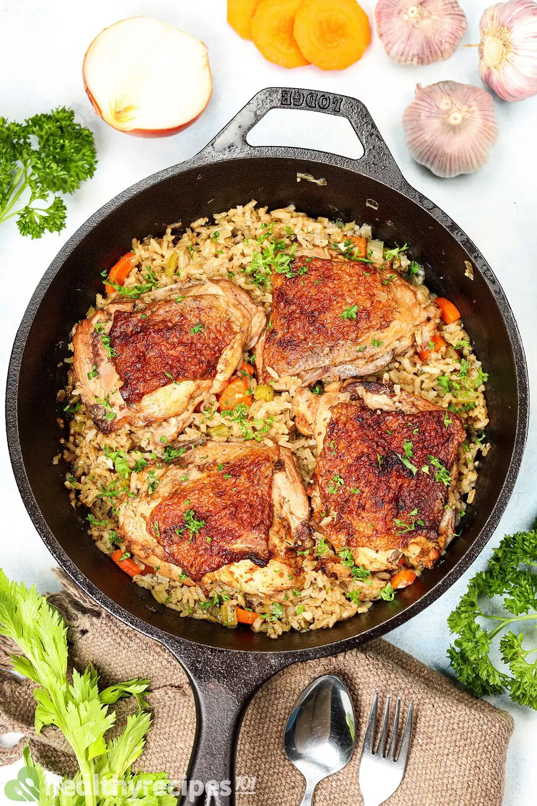Four chicken thighs on top of cooked rice in a cast iron skillet decorated with a tablecloth, utensils, and ingredients of the recipe