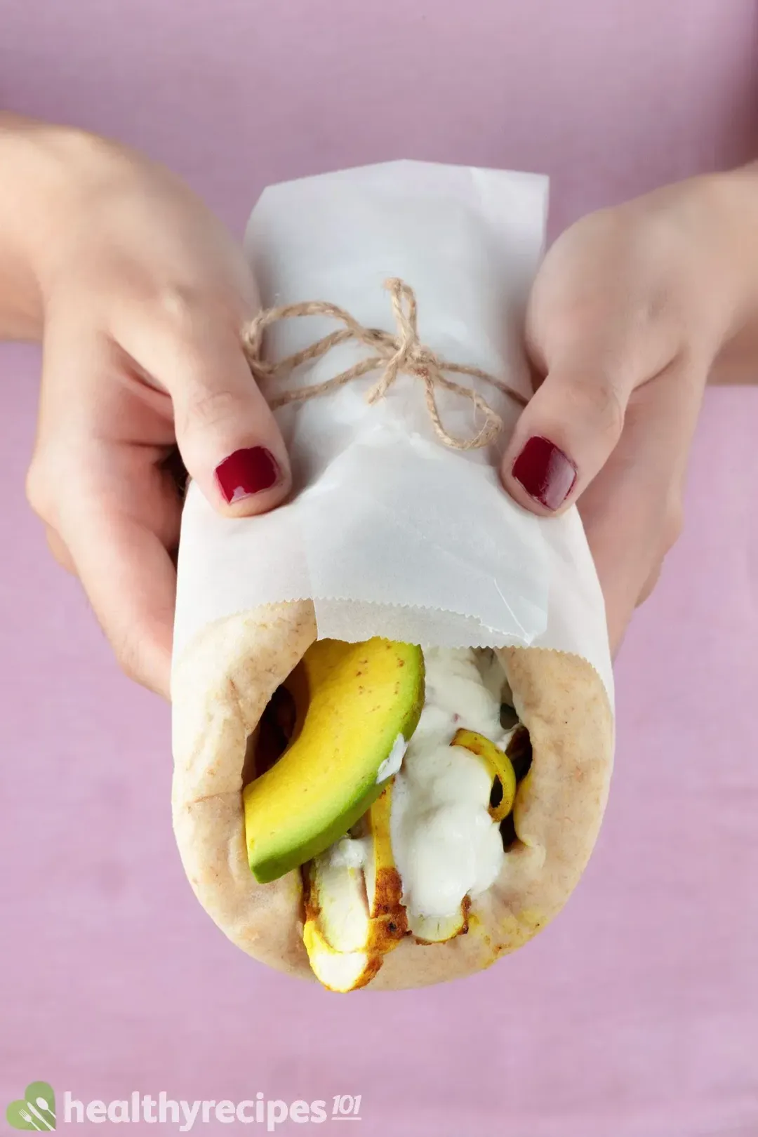 Two hands presenting a chicken shawarma wrapped in parchment and tied with brown twine