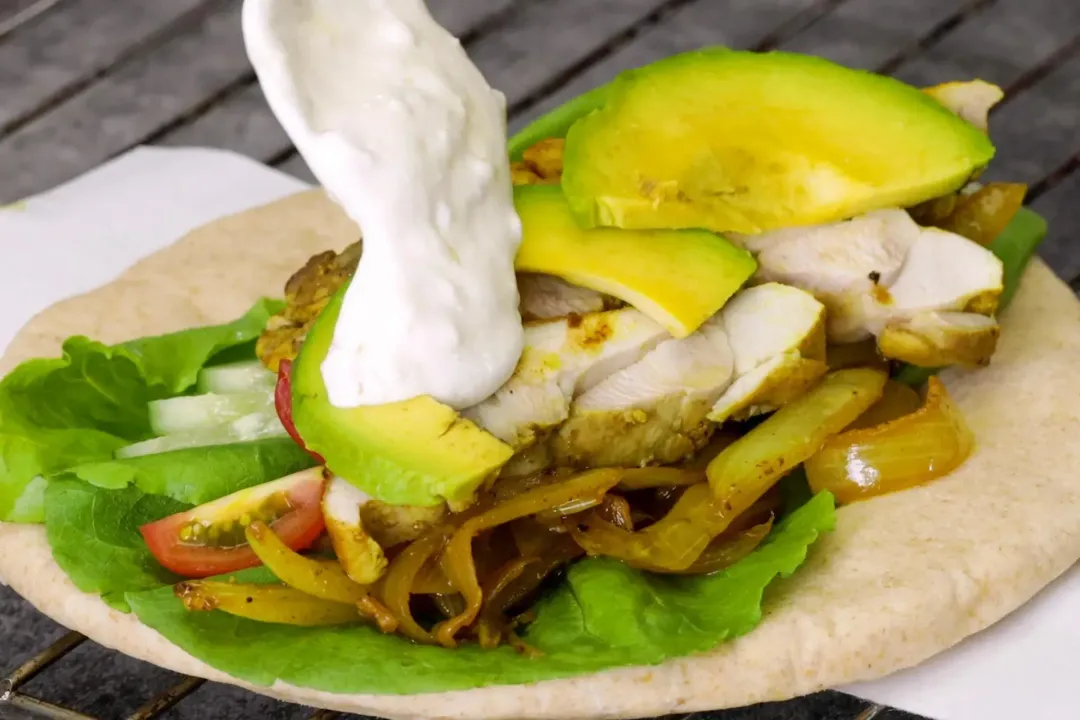 A pita bread topped with vegetables, avocado slices, and cooked chicken dolloped with white sauce