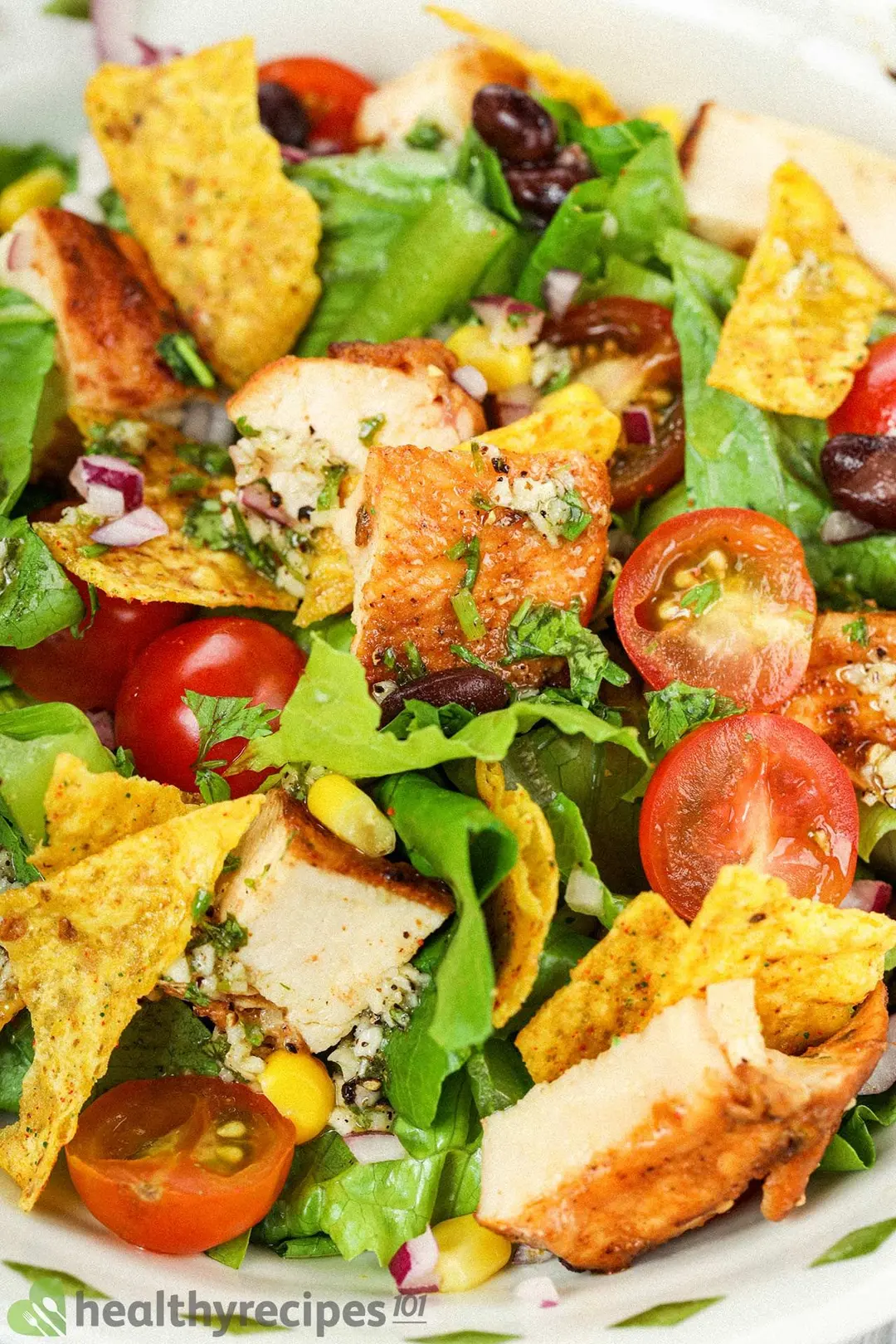 A close up on a chicken taco salad filled with cooked chicken pieces, dorioto chips, shredded lettuce, halved cherry tomatoes, and corn