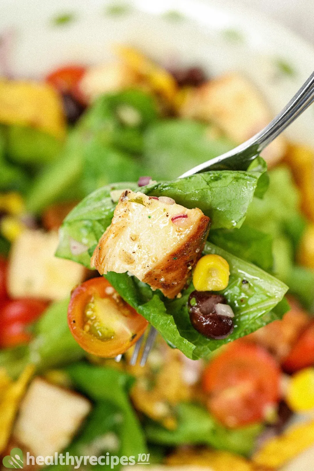 A fork piercing through lettuce pieces, half a cherry tomato, a cooked chicken cube, and a black bean