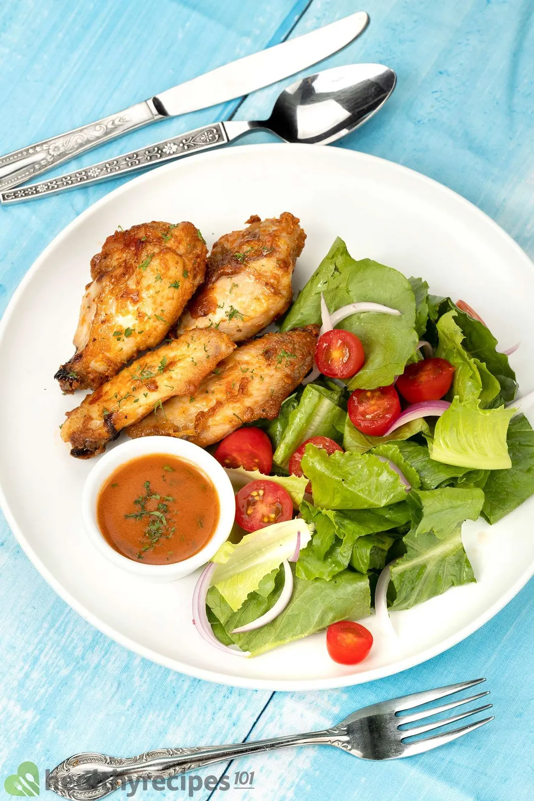 a white plate of cooked chicken wings with half tomatoes and chopped lettuce with a small bowl of sauce, decorated with a fork, knife and spoon