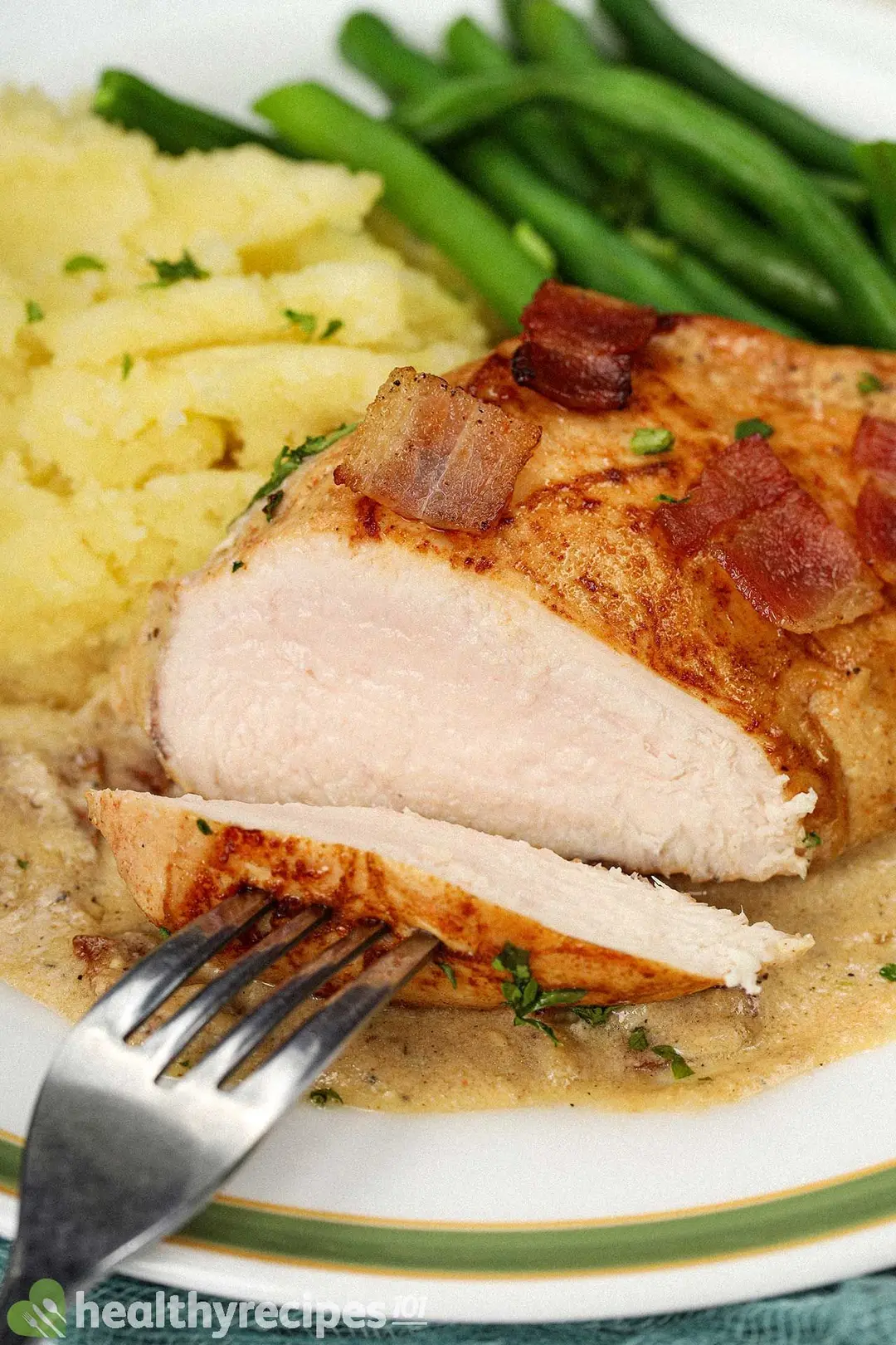 A fork sticking into a piece of chicken with another large piece of chicken, mashed potatoes, bacon pieces, and green beans in the background