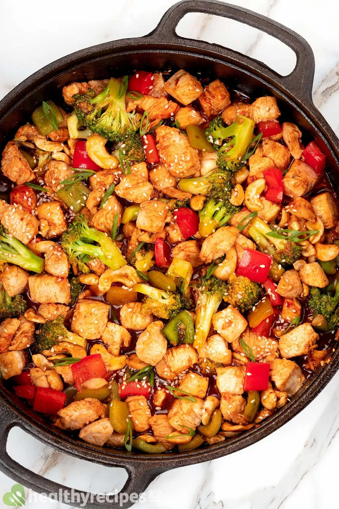 a cast iron skillet of cooked cubed chicken with cubed pepper, broccoli and cashew decorated with sesame seeds and chopped scallion