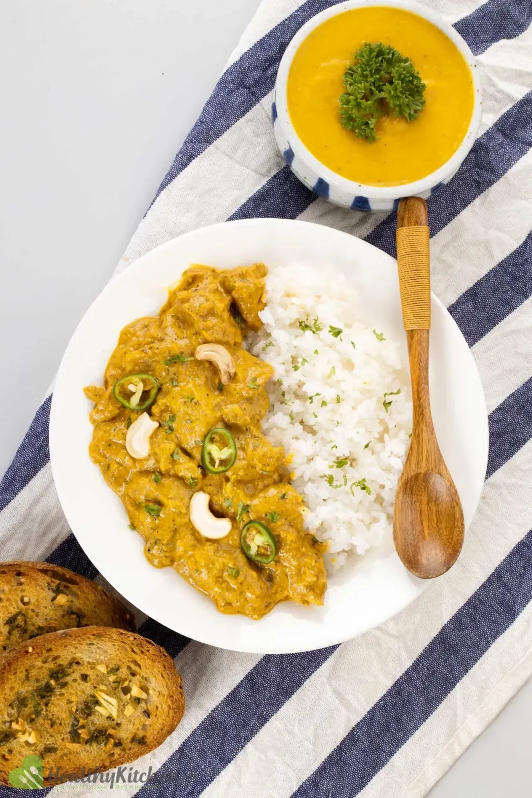 A plate of chicken korma and cooked rice with a wooden spoon on top, next to garlic bread and pumpkin soup