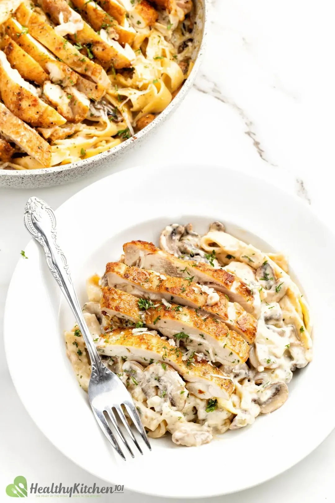 A plate of cooked chicken thigh next to a chicken thigh skillet, full of sliced grilled chicken, mushrooms, pasta, and white mushroom sauce