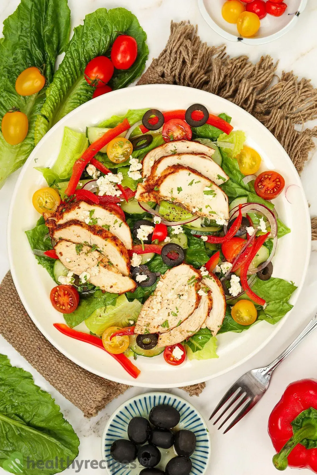 A plate of Greek Chicken Salad placed on a brown cloth and near lettuce leaves, black olives, and cherry tomatoes.