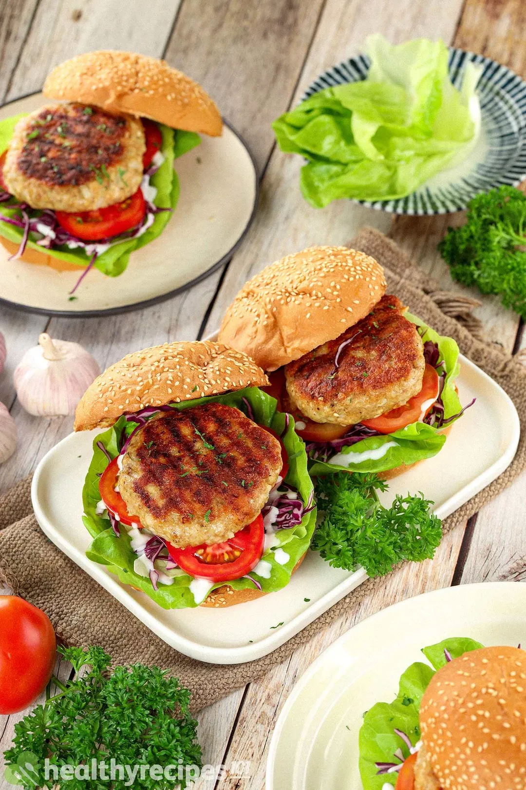 Plates of burgers containing meat patties, tomatoes, shredded cabbage, and lettuce, decorated by parsley and a brown fabric