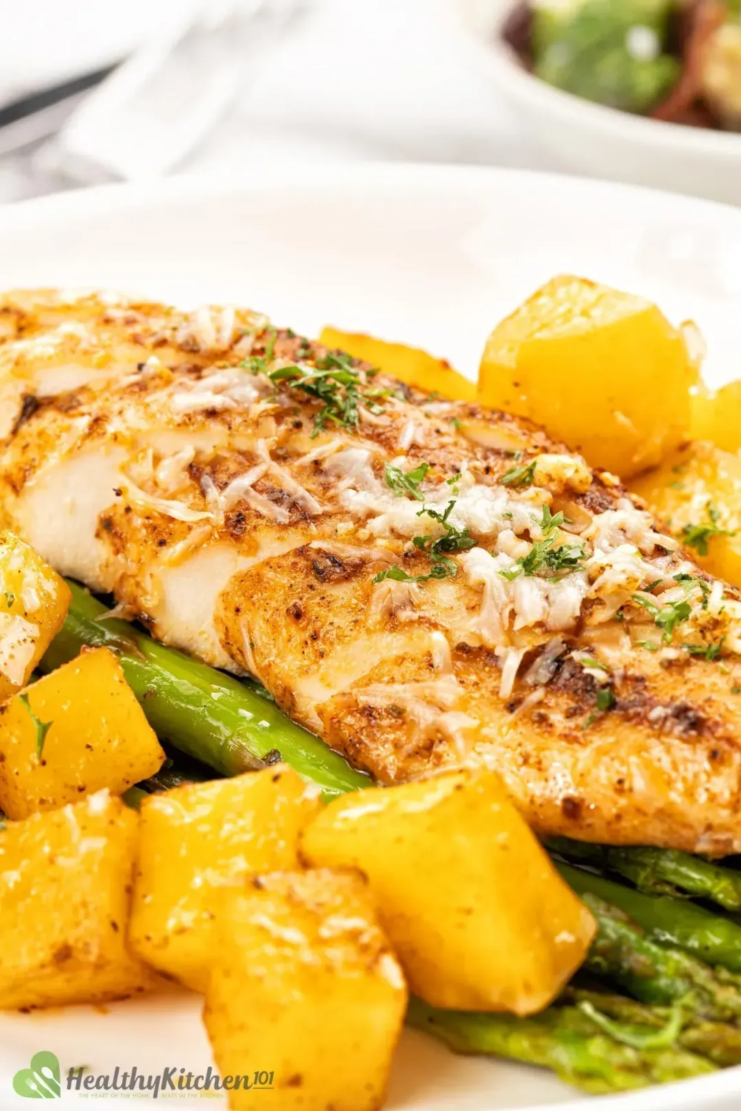 A grilled chicken breast coated in seasonings and topped with grated parm, next to baked potato cubes and on buttered asparagus